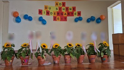 Acht Blumentöpfe mit Sonnenblume stehen auf einer Bühne. An der Wand dahinter sind blaue und orangene Luftballons aufgehängt, dazwischen ein Spruch: Ade du schöne Kindergartenzeit